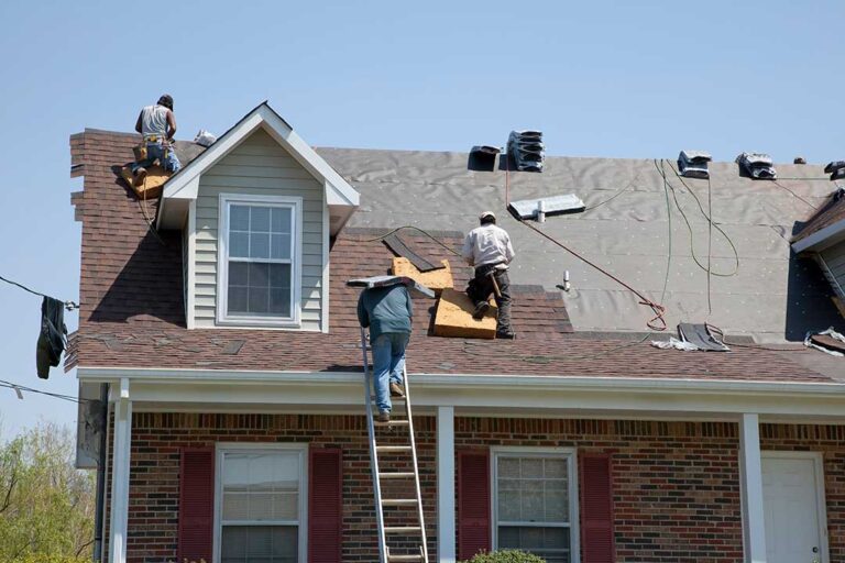 A roofer removing old shingles from a house roof with plywood decking exposed.