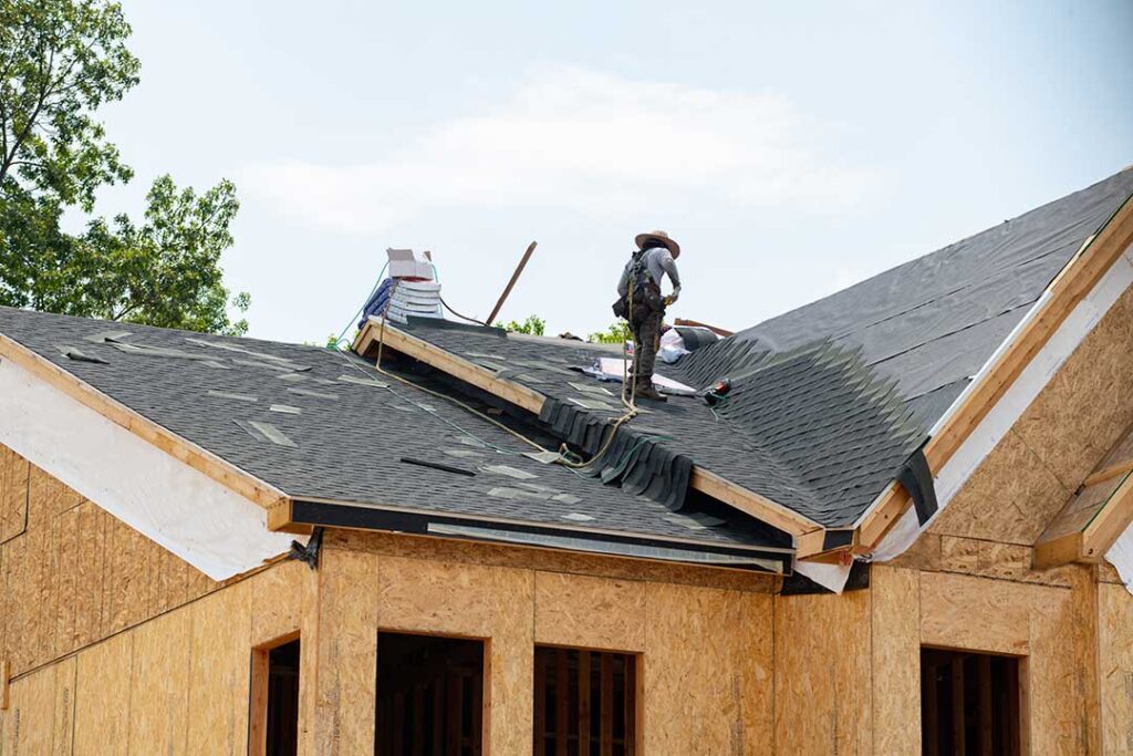A homeowner inspects gutters beside a residential roof while autumn leaves gather below.